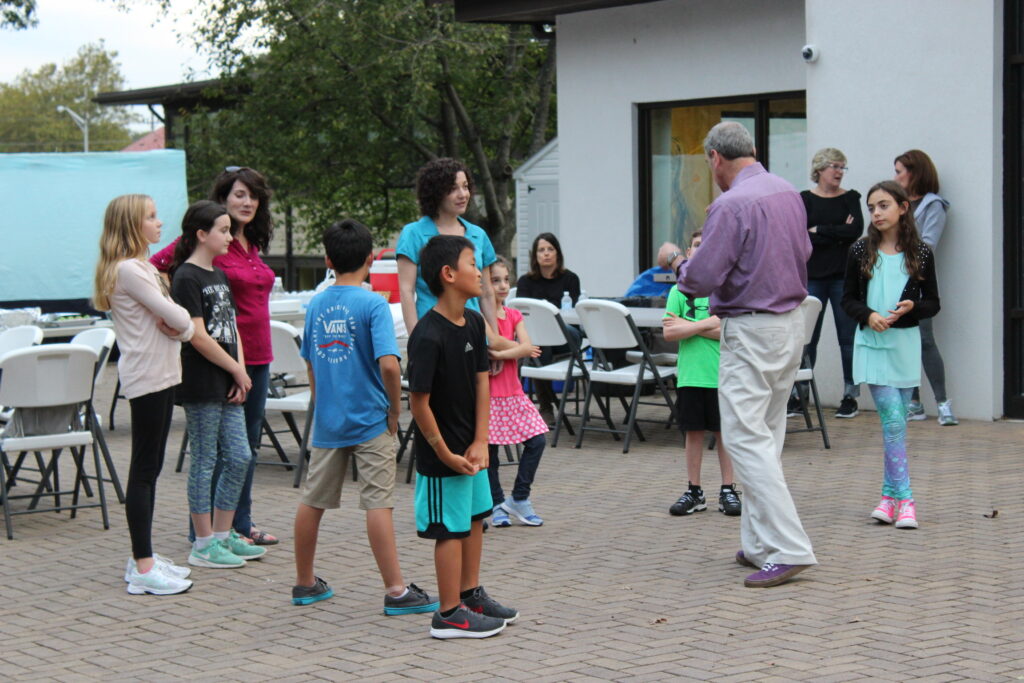 Group of children and adults gathered outdoors, listening to a man speaking.