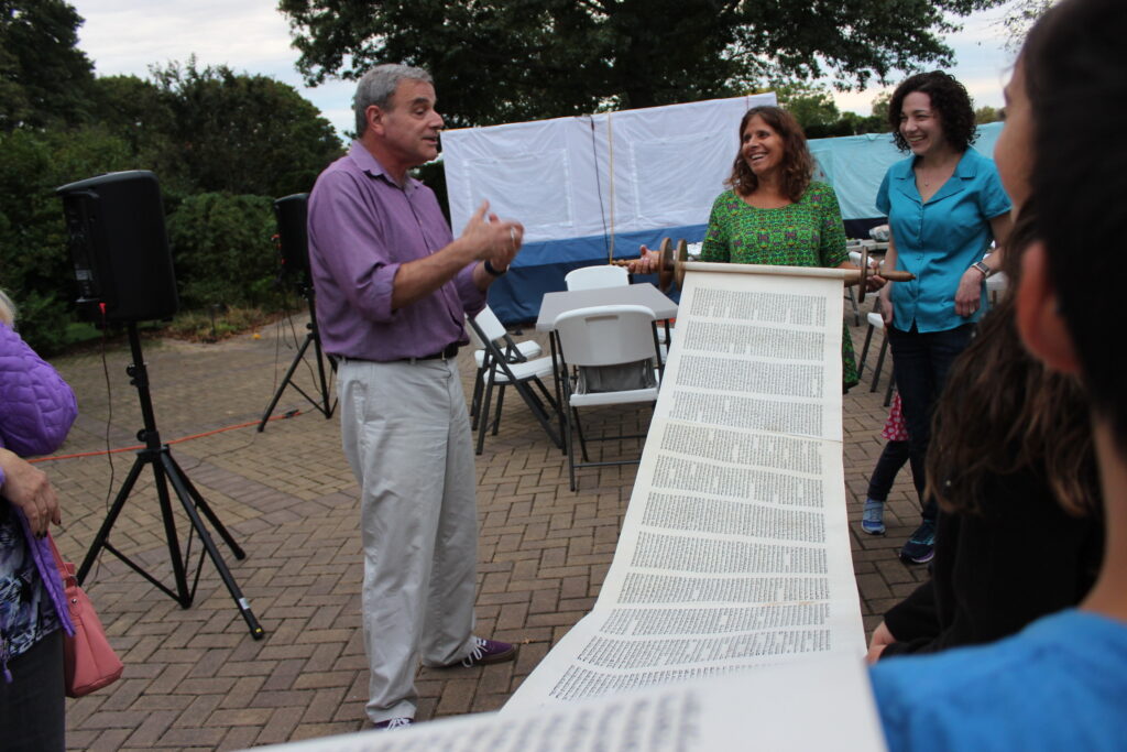 Man reading from an extremely long scroll of paper outdoors with two women.