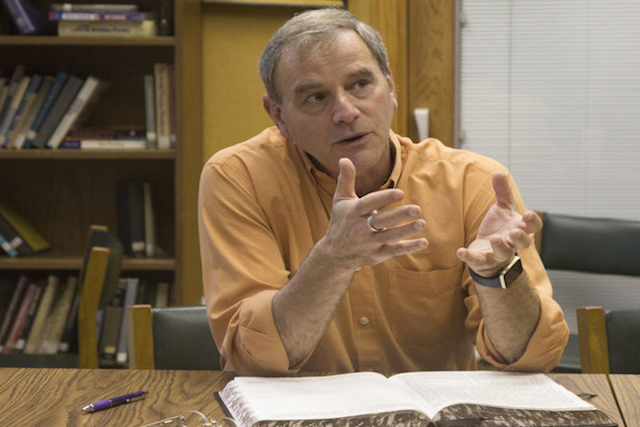 Man in orange shirt gestures while seated at desk with open book.