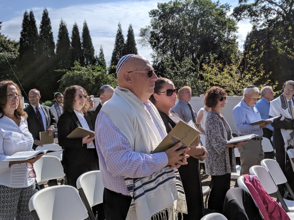 People gathered outdoors, some holding books, in a sunny setting.