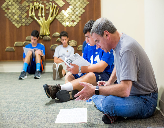 Man teaching a group of kids seated on the floor with papers.