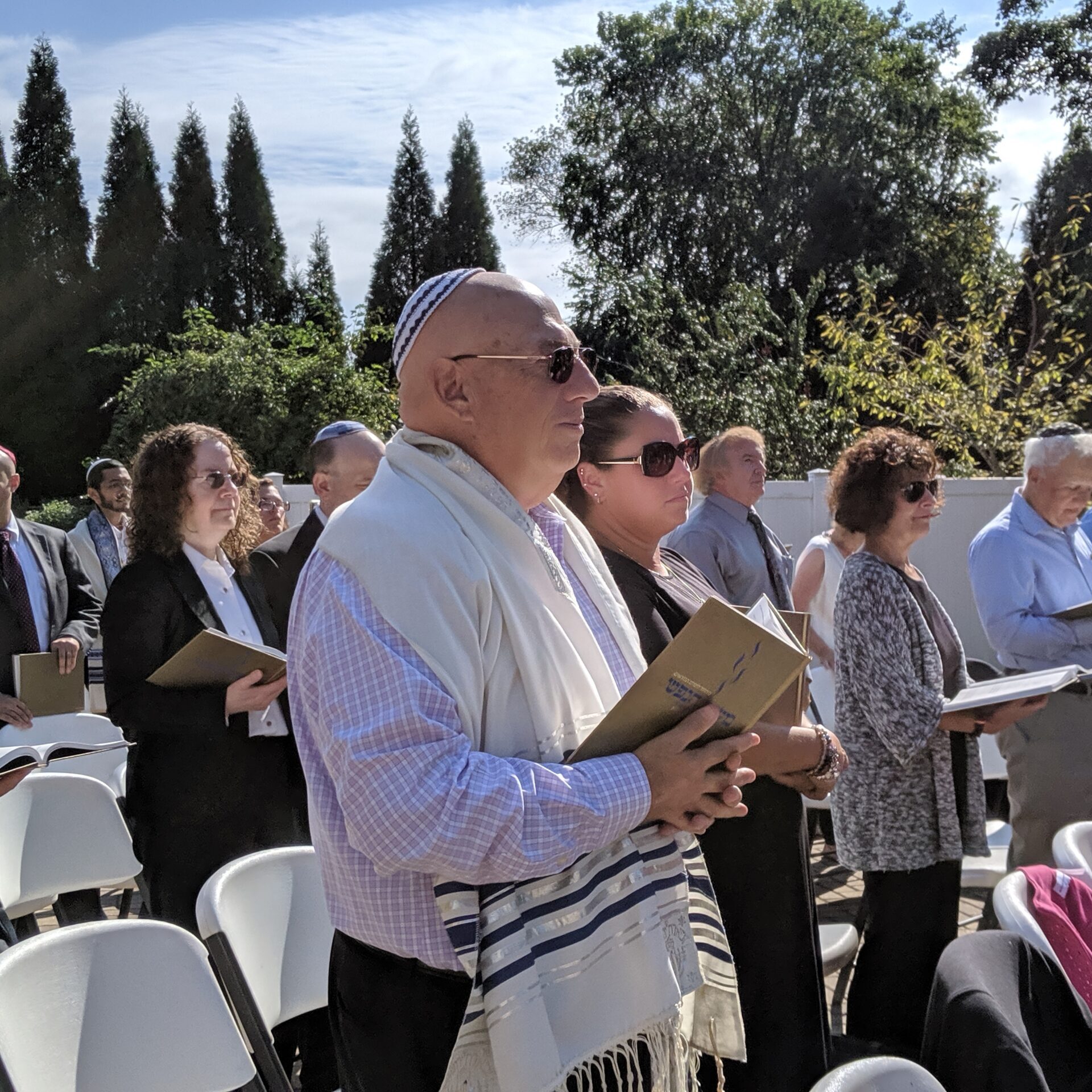 People gathered outdoors, some holding books, in a sunny setting.