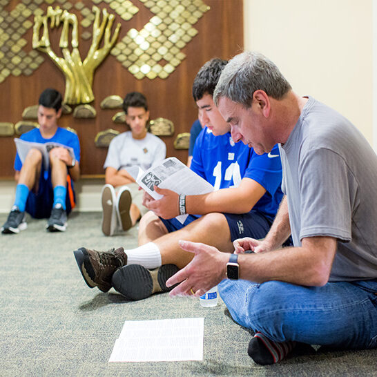 Man teaching a group of kids seated on the floor with papers.