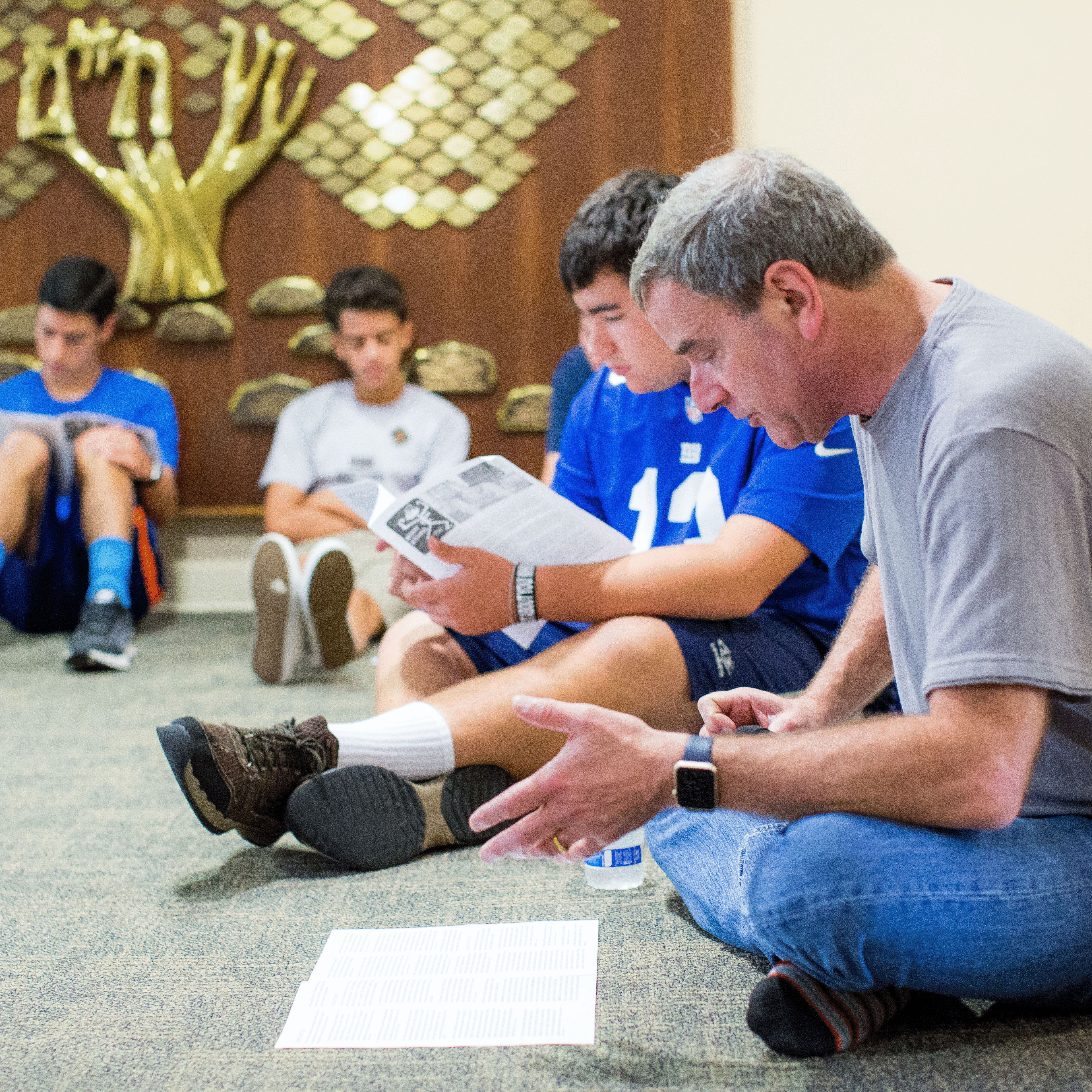 Group of men sitting on floor reading papers in a casual setting.