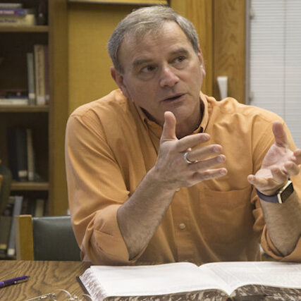 Man in orange shirt gestures while seated at desk with open book.