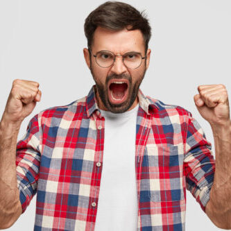 Furious angry man with irritated expression, clutches fists angrily, shouts at someone, wears checkered shirt, stands against white background. Negative human emotions and feelings. Body language Furious angry man with irritated expression, clutches fists angrily, shouts at someone, wears checkered shirt, stands against white background. Negative human emotions and feelings. Body language